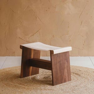 May include: A handcrafted wooden stool with a dark brown frame and a white fabric seat. The stool is made of solid wood and has a simple, modern design. It is placed on a round, woven rug.