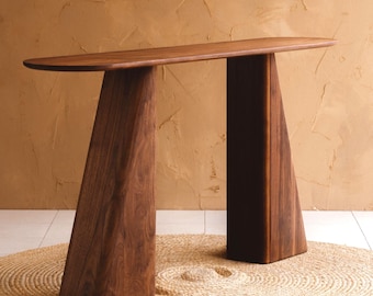 Può includere: Un tavolo console in legno marrone scuro caratterizzato da una lunga superficie superiore liscia e ovale, sostenuta da due gambe larghe, affusolate e trapezoidali. Il mobile mostra un motivo a venature del legno naturale in tutta la sua solida struttura.