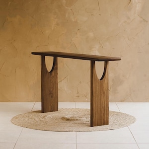 May include: A dark wood console table with a rectangular top and two vertical supports. Each support has a curved cutout. The table is set on a round, woven jute rug. The table's dimensions are approximately 60 inches long, 12 inches wide, and 30 inches high.