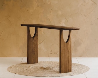 Può includere: Un tavolo console in legno marrone scuro con un piano rettangolare lungo e stretto sostenuto da due gambe verticali. Ogni gamba presenta un intaglio a forma di U vicino alla parte superiore, creando un design ad arco aperto sotto il piano.