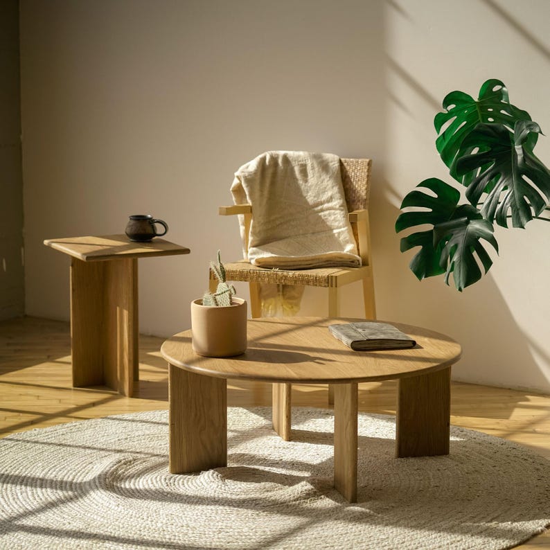 May include: A light-filled living space with a round wooden coffee table and a small side table. A woven chair holds a beige blanket. A small potted plant and a mug are on the tables. A monstera plant adds greenery to the scene.
