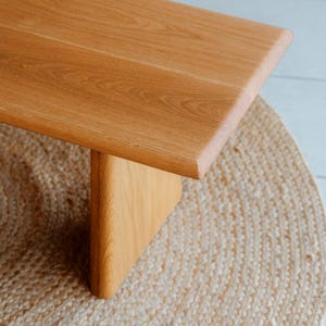 Puede incluir: Un banco de madera de color marr&oacute;n claro con una superficie rectangular lisa de bordes redondeados y una pata de soporte vertical s&oacute;lida. La veta de la madera es visible en toda la superficie, mostrando patrones naturales en un tono miel.