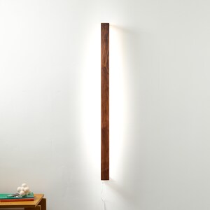 Puede incluir: Aplique de pared rectangular de madera con un acabado marr&oacute;n c&aacute;lido. La l&aacute;mpara tiene una fuente de luz lineal y brillante que la atraviesa. Est&aacute; montada en una pared blanca, con un cable de alimentaci&oacute;n visible en la parte inferior.