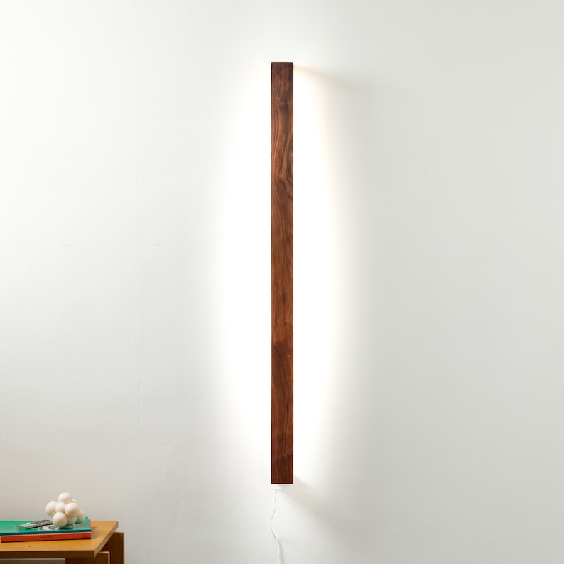Puede incluir: Aplique de pared rectangular de madera con un acabado marr&oacute;n c&aacute;lido. La l&aacute;mpara tiene una fuente de luz lineal y brillante que la atraviesa. Est&aacute; montada en una pared blanca, con un cable de alimentaci&oacute;n visible en la parte inferior.