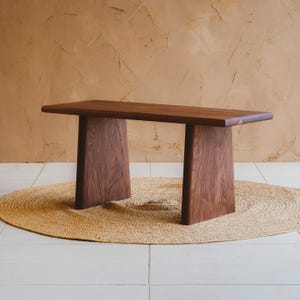 Puede incluir: Un banco rectangular de madera con un acabado de grano marr&oacute;n oscuro y liso. El banco est&aacute; construido con una superficie superior horizontal plana sostenida por dos patas verticales anchas y s&oacute;lidas que se estrechan ligeramente hacia arriba.
