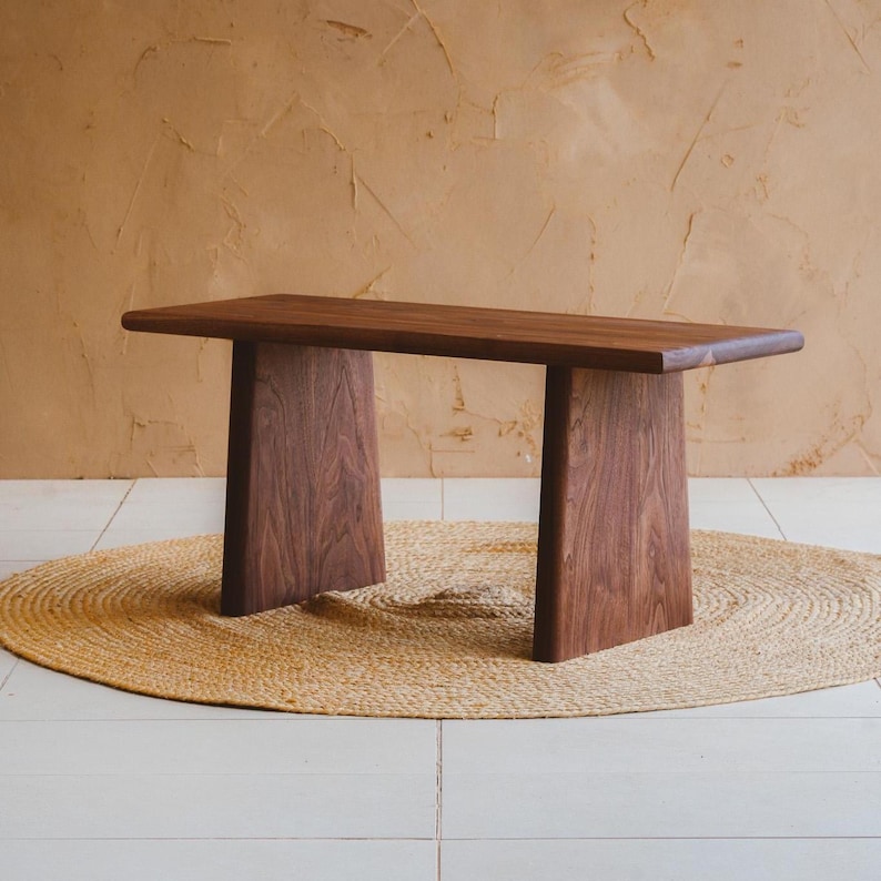 Puede incluir: Un banco rectangular de madera con un acabado de grano marr&oacute;n oscuro y liso. El banco est&aacute; construido con una superficie superior horizontal plana sostenida por dos patas verticales anchas y s&oacute;lidas que se estrechan ligeramente hacia arriba.
