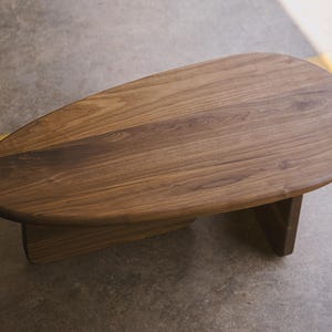 May include: A low, oval-shaped wooden coffee table. The table has a smooth, dark brown surface with visible wood grain. It rests on two rectangular supports, creating a minimalist design. The table is approximately 48 inches long.