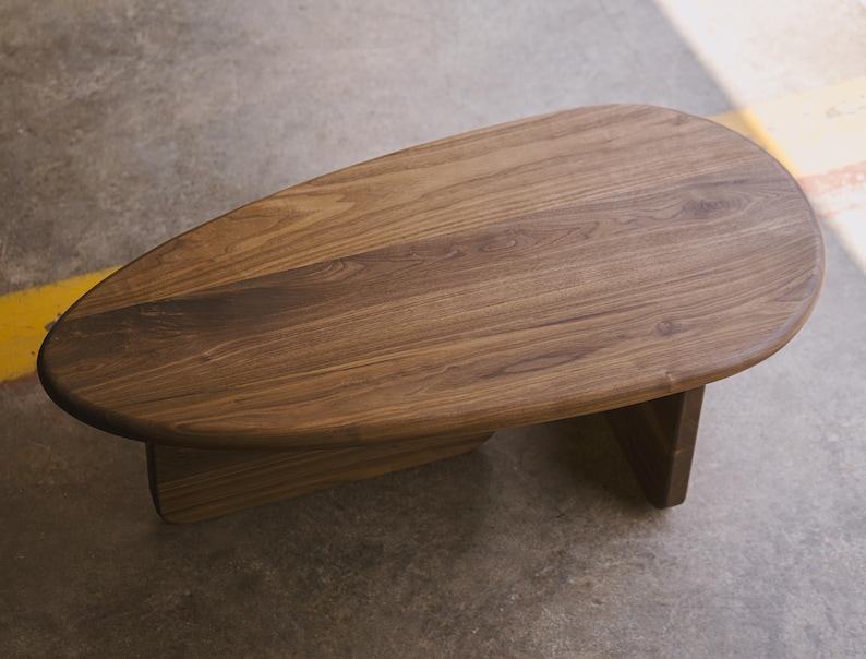 May include: A low, oval-shaped wooden coffee table. The table has a smooth, dark brown surface with visible wood grain. It rests on two rectangular supports, creating a minimalist design. The table is approximately 48 inches long.