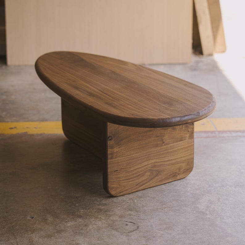 May include: A solid wood coffee table with a smooth, oval top and a rectangular base. The table is made of a warm, medium-toned wood, showcasing the natural grain. The design is minimalist and modern, suitable for a living room.