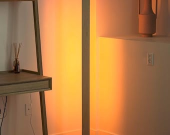 May include: A modern wooden floor lamp with a rectangular design. The lamp has a light wood finish and emits a warm, orange glow. The lamp is standing on a rectangular base and is placed in the corner of a room.
