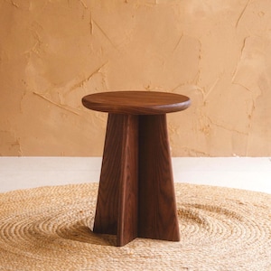 May include: A dark brown wooden stool with a round top and a triangular base. The stool is made of solid wood and has a natural finish. It is placed on a round, woven jute rug against a textured, tan wall.