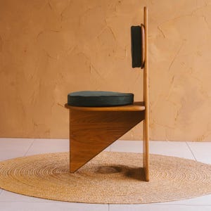 K&ouml;nnte beinhalten: Ein Holzstuhl mit einer hohen, flachen R&uuml;ckenlehne und einem dreieckigen Untergestell. Der Stuhl verf&uuml;gt &uuml;ber ein dunkles, rundes Sitzkissen und ein passendes dunkles, rechteckiges R&uuml;ckenpolster an der vertikalen Holzplatte. Das Holz ist naturfarben.