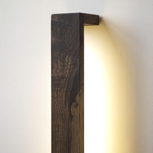 Puede incluir: Aplique de pared rectangular de madera marr&oacute;n oscuro con una luz c&aacute;lida y difusa. La veta de la madera es visible, a&ntilde;adiendo textura. El aplique est&aacute; montado en una pared blanca, creando un contraste.