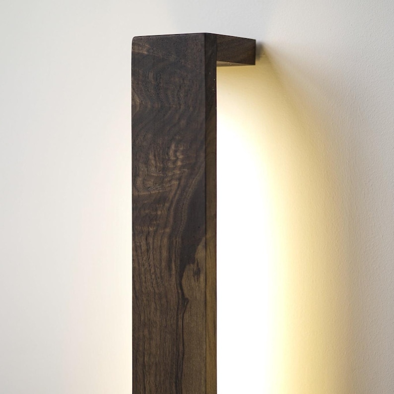 Puede incluir: Aplique de pared rectangular de madera marr&oacute;n oscuro con una luz c&aacute;lida y difusa. La veta de la madera es visible, a&ntilde;adiendo textura. El aplique est&aacute; montado en una pared blanca, creando un contraste.