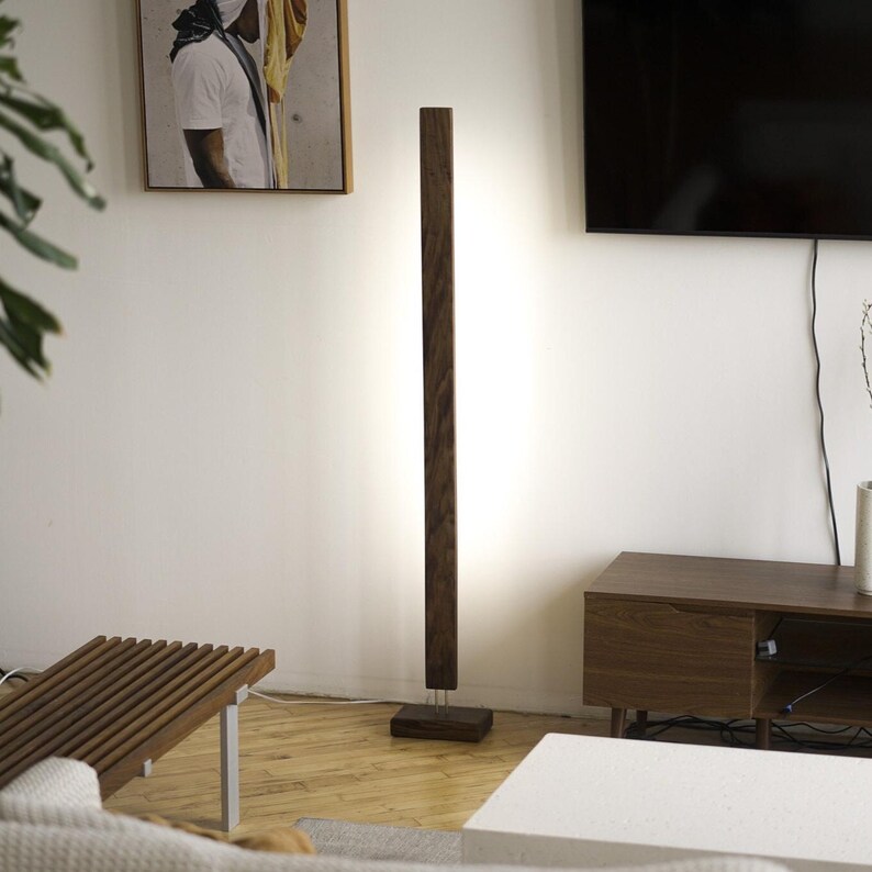 May include: A modern, dark wood floor lamp with a rectangular base and a tall, vertical light column. The lamp is positioned in a living room setting, providing ambient lighting. The lamp's height is approximately 60 inches.