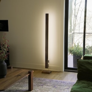 May include: A modern, minimalist floor lamp with a tall, rectangular wooden frame. The lamp emits a soft, warm light, creating a vertical illuminated strip. The base is a dark wood, and the lamp is placed in a living room setting.