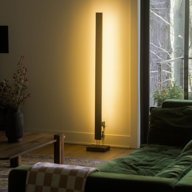 May include: A modern, minimalist floor lamp with a tall, rectangular wooden frame. The lamp emits a warm, inviting light, creating a vertical stripe of illumination against the wall. The lamp is set on a dark wooden base.
