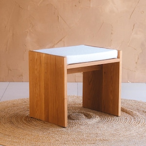 May include: A rectangular wooden stool featuring a light brown natural wood grain finish and a thick, flat white upholstered seat cushion. The stool has two solid side panels that serve as legs, providing a stable base for the rectangular top.