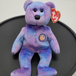 May include: A plush teddy bear with a tie-dye pattern of purple and blue. The bear features black plastic eyes and a black nose, a purple ribbon around its neck, and a Ty Beanie Babies heart-shaped tag attached to its ear. A circular Ty Official Club patch is on its chest.