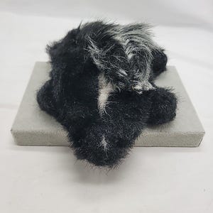 May include: A small, plush skunk figurine covered in black and white faux fur. The skunk features a prominent white stripe running from its head down its back, with a textured, fuzzy appearance. It is positioned in a resting, prone posture.