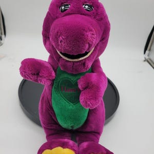 Puede incluir: Un peluche de dinosaurio morado con un parche verde en el vientre en forma de corazón y el texto I Love You. El juguete tiene grandes ojos negros, una amplia sonrisa y pies amarillos. Se muestra en posición sentada con los brazos extendidos.