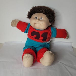 以下が含まれることがあります： 柔らかい体と丸い顔が特徴の、ヴィンテージのキャベツ人形。 赤とティールのフットボールのユニフォームを着ており、背番号は21です。 茶色のふわふわした髪で、腕を広げてポーズをとっています。