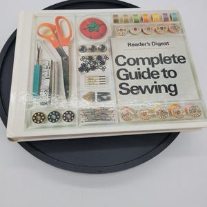 Op de afbeelding: Het boek Reader's Digest Complete Guide to Sewing heeft een witte kaft met een rasterindeling die naaigereedschap toont, waaronder een schaar met oranje handvatten, een tornmesje, een meetlint, klossen garen, naalden en knopen.