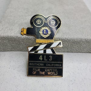 May include: A vintage film camera pin with a clapperboard base. The pin is black, gold, and white, with the text "4 L 3", "Southern California", and "Film Capital of the World". The camera has a Lions Club logo.