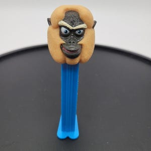 May include: A Pez candy dispenser featuring a molded plastic head of a primate character with dark gray facial features, light brown fur, and blue eyes, attached to a vertical blue plastic stem with a grooved design and a flared base.