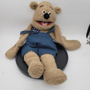 May include: A tan plush bear puppet with a fuzzy texture, large white eyes with black pupils, and a black nose. The bear has an open mouth with a red interior and a white tongue. It wears blue denim overalls with a blue and white polka dot bowtie.