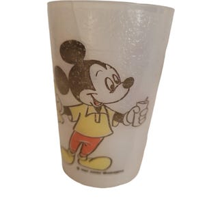 May include: A clear plastic cup featuring a smiling Mickey Mouse cartoon character. Mickey is wearing a yellow shirt, red pants, and yellow shoes. The cup has a slightly textured surface and a tapered shape. The image is a vintage design.