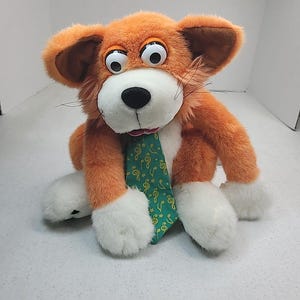 Puede incluir: Un peluche de animal naranja y blanco con grandes ojos móviles, nariz negra y bigotes largos. El juguete lleva una corbata verde decorada con un patrón de notas musicales y estrellas amarillas, con patas blancas y orejas grandes y caídas.