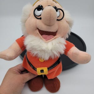 Puede incluir: Un peluche del personaje Doc de Blancanieves y los siete enanitos, con cara color canela, barba blanca, túnica naranja, cinturón negro con hebilla amarilla y gafas negras redondas. El personaje tiene grandes ojos blancos y una amplia sonrisa.