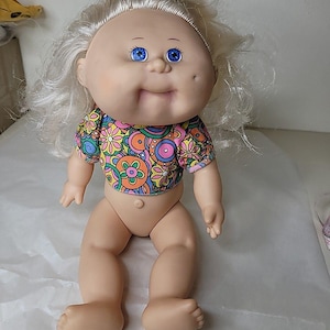 May include: A vintage doll with a round face, blue eyes, and blonde hair. The doll is wearing a colourful floral print shirt. The doll is sitting with its legs spread apart.