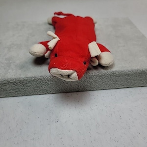 May include: A red plush toy cow with white detailing on its hooves, ears, and snout. The toy is positioned on a grey surface, highlighting its soft, textured fabric. The cow features small black eyes and a simple design.