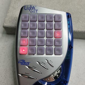 Può includere: Gioco elettronico portatile "Lights Out" di Tiger Games. Custodia in plastica argento e blu con una griglia di pulsanti viola, alcuni illuminati di rosso. Pulsanti "ON/OFF", "START", "SOUND" e "HELP".