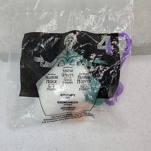 May include: A sealed plastic bag containing a Grumpy toy from the Disney movie Snow White and the Seven Dwarfs. The packaging features the character Grumpy and text in multiple languages, including English, French, and Spanish. A purple plastic piece is attached.