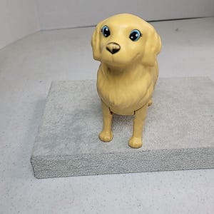 May include: A tan-colored plastic toy dog with blue eyes. The dog is standing on a gray surface. The toy dog has a friendly expression and is a miniature replica of a Golden Retriever.