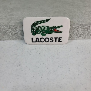 Op de afbeelding: Wit rechthoekig embleem met een groene krokodil en rode mond. Het woord "LACOSTE" staat eronder in het zwart gedrukt. Een klassiek ontwerp.