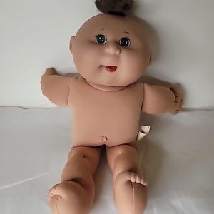 May include: A vintage Cabbage Patch Kid doll with a bald head, green eyes, and a small tuft of brown hair. The doll has a peach-coloured skin tone, a red mouth, and is wearing no clothing. The doll is in a sitting position with arms outstretched.