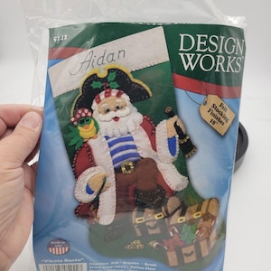 May include: A Design Works felt stocking craft kit titled Pirate Santa, featuring a Santa Claus dressed as a pirate with a parrot, treasure chest, and the name Aidan printed at the top. The packaging indicates the finished stocking is 18 inches.