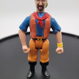 May include: A plastic action figure of a pirate with a green headscarf, a blue shirt, a red neckerchief, an orange vest, and orange pants. The figure has a black goatee, black boots, and two yellow bracelets on one wrist, with a black belt buckle.