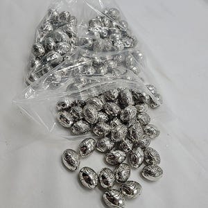 Puede incluir: Una colección de cuentas plateadas de forma ovalada con intrincados patrones florales en relieve. Las cuentas están contenidas en una bolsa de plástico transparente, con varias cuentas individuales derramadas sobre la superficie.
