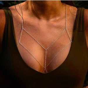 Puede incluir: Una delicada cadena de cuerpo dorada. La cadena está diseñada para cubrir el pecho y el torso, creando un patrón geométrico. La cadena se usa sobre una camiseta verde oscuro.