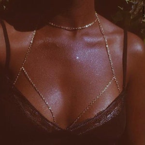 May include: A gold-colored body chain necklace. The necklace features a delicate chain that drapes across the chest and connects to a choker-style necklace. The model is wearing a black lace top.