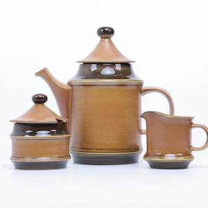 Pode incluir: Um conjunto de chá de cerâmica marrom e preto com bule, jarra de creme e açucareiro. O conjunto tem um design simples e elegante com um esmalte marrom e detalhes pretos.