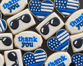 May include: Assortment of decorated sugar cookies with blue and white icing. Some cookies feature the American flag with a blue line, others have sunglasses, and some say "thank you".