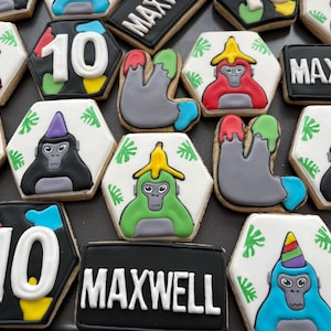 May include: Assortment of decorated sugar cookies. Some cookies are hexagon-shaped with a white background and a cartoon monkey design. Other cookies are rectangular with the name "MAXWELL" or the number "10". The cookies are decorated with various colors.