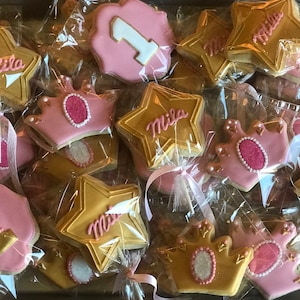May include: A collection of decorated sugar cookies in pink and gold. The cookies are shaped like stars and crowns and feature the name "Mila" and the number "1".