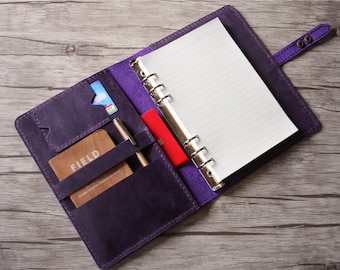 Op de afbeelding: Een paarse lederen planner met een zilveren ringband. De planner is open en laat een blanco, gestreept blad zien. De planner heeft een zakje op de voorzijde en een pennenhouder aan de zijkant. De planner ligt op een houten oppervlak.